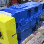 Large industrial gearbox freshly powder coated yellow and blue sitting on wooden pallet in repair facility