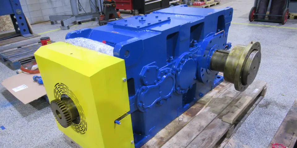 Large industrial gearbox freshly powder coated yellow and blue sitting on wooden pallet in repair facility